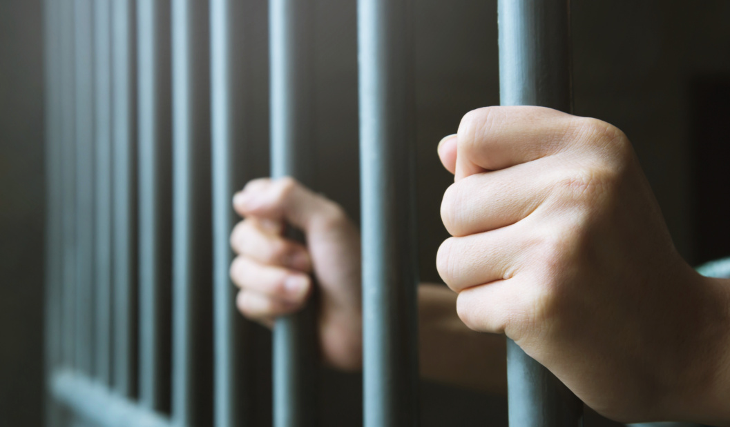 Man In Prison Hands Of Behind Hold Steel Cage Jail Bars. Offender Criminal Locked In Jail.